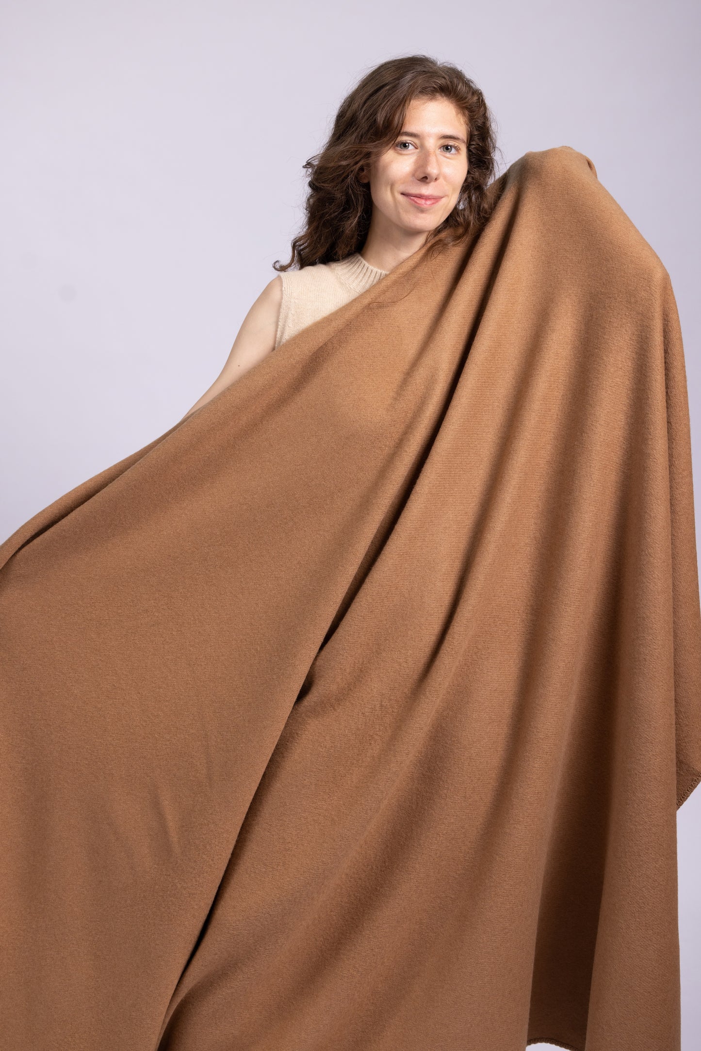 This image highlights a camel wool blanket being held and displayed, showcasing its large size and elegant drape. The blanket's warm camel hue exudes a natural and sophisticated aesthetic. The fabric appears soft, smooth, and lightweight, likely made from high-quality wool. Its generous proportions and flowing design make it versatile for use as a cozy throw, wrap, or home accessory. The blanket's craftsmanship is apparent in its seamless finish, emphasizing comfort, luxury, and timeless style.