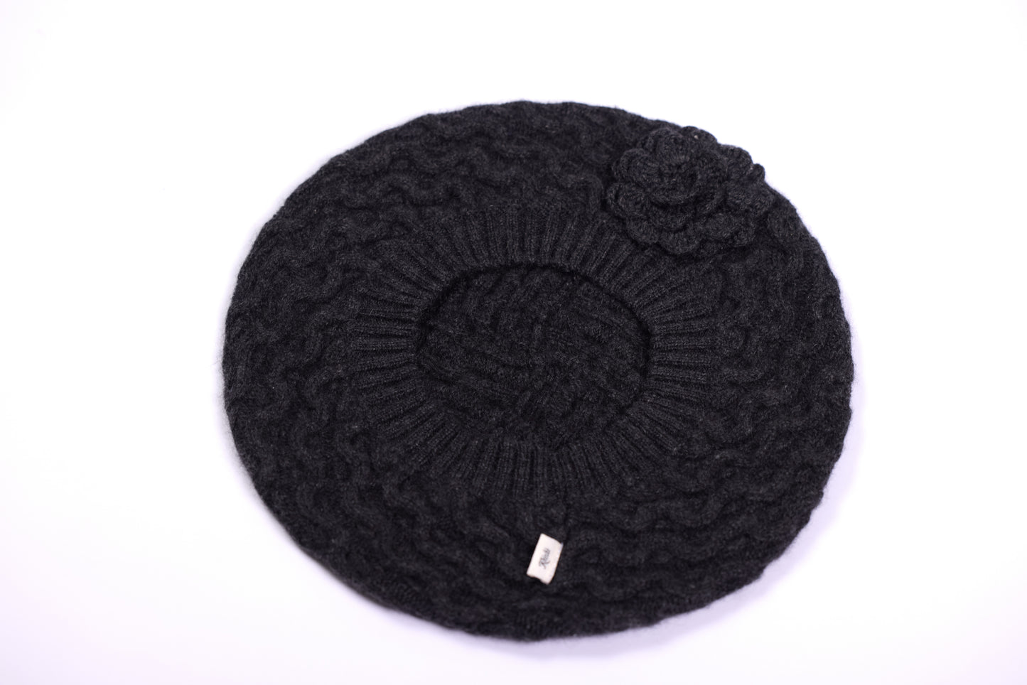 This is a luxurious cashmere beret in a dark grey color. It features a classic cable-knit design that adds texture and dimension. The beret is adorned with a delicate, handcrafted flower accent on the side, enhancing its timeless elegance. The ribbed edge provides a snug yet comfortable fit, making it a chic and cozy accessory for cooler seasons.