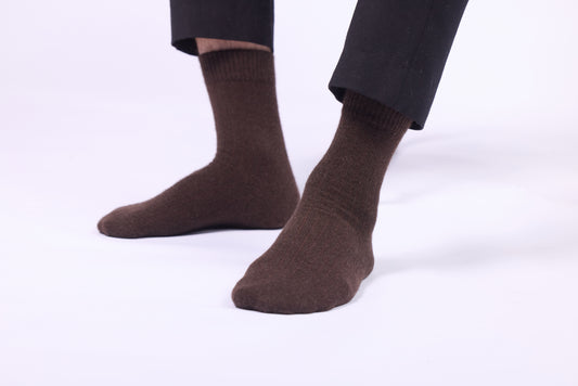 These dark brown yak wool socks are displayed on a person standing with bare legs, highlighting the socks' height and snug fit. The socks rise to a mid-calf length, featuring a ribbed cuff at the top to keep them securely in place. The texture appears soft and dense, with a smooth and uniform knit, reflecting the natural insulating and plush qualities of yak wool. These socks functionality, style, and luxury, making them suitable for casual or cozy indoor wear.