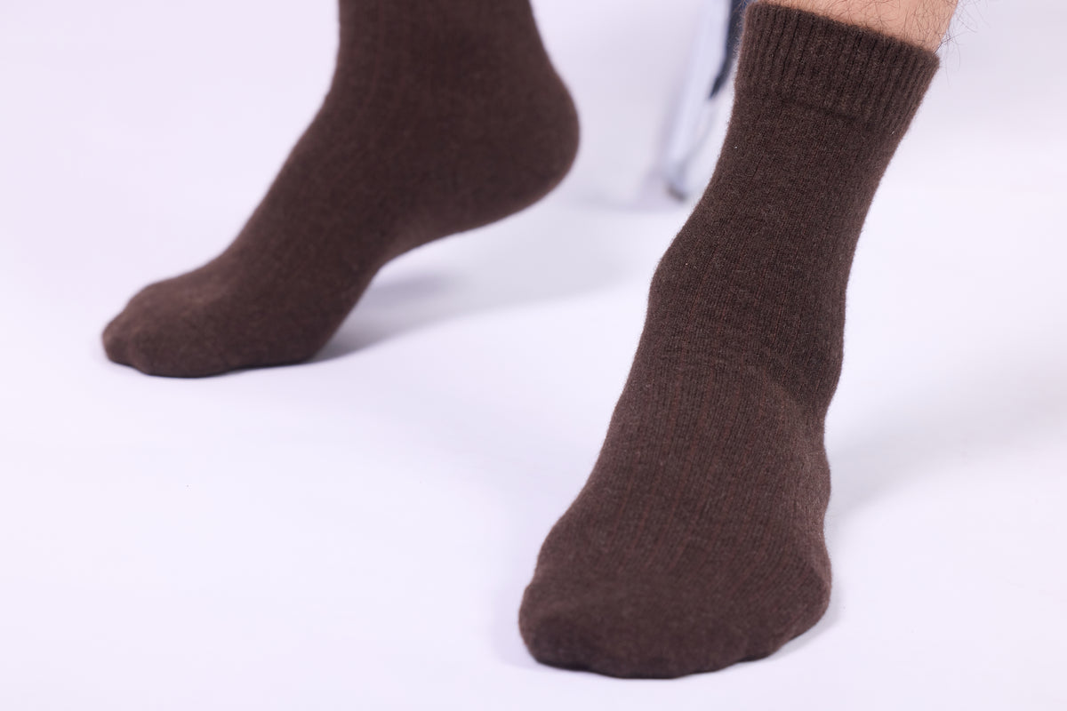These dark brown yak wool socks are showcased in close-up, emphasizing their snug fit and cozy appearance. The socks have a smooth, slightly ribbed texture, extending up to the lower calf. The natural dark brown tone gives them an elegant and earthy aesthetic. The thickness and plush quality of the socks suggest superior warmth and comfort, ideal for colder climates. Yak wool, known for its softness, durability, and insulation, makes these socks a luxurious and practical choice for daily wear. 